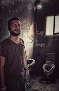 A man stands in a dimly lit, deteriorating bathroom with dark, stained walls and floors. The room appears worn with visible grime and decay. A damaged sink and toilet are present with a small, dirty window letting in minimal light.