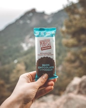 A hand holding a snack bar labeled 'Perfect Snacks' with 'Dark Chocolate Coconut' flavor, set against an outdoor backdrop featuring blurred mountains and trees.
