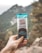 A hand holding a snack bar labeled 'Perfect Snacks' with 'Dark Chocolate Coconut' flavor, set against an outdoor backdrop featuring blurred mountains and trees.
