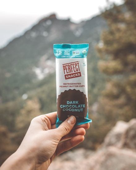 A hand holding a snack bar labeled 'Perfect Snacks' with 'Dark Chocolate Coconut' flavor, set against an outdoor backdrop featuring blurred mountains and trees.