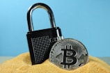 A metallic padlock is partially buried in sand next to a physical representation of a Bitcoin coin. The backdrop is a solid light blue, creating a contrast with the golden sand and the metallic objects.