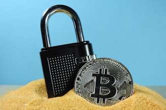 A metallic padlock is partially buried in sand next to a physical representation of a Bitcoin coin. The backdrop is a solid light blue, creating a contrast with the golden sand and the metallic objects.