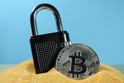A metallic padlock is partially buried in sand next to a physical representation of a Bitcoin coin. The backdrop is a solid light blue, creating a contrast with the golden sand and the metallic objects.