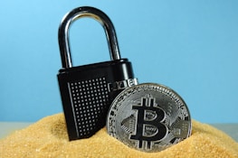 A metallic padlock is partially buried in sand next to a physical representation of a Bitcoin coin. The backdrop is a solid light blue, creating a contrast with the golden sand and the metallic objects.