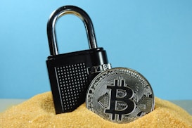 A metallic padlock is partially buried in sand next to a physical representation of a Bitcoin coin. The backdrop is a solid light blue, creating a contrast with the golden sand and the metallic objects.