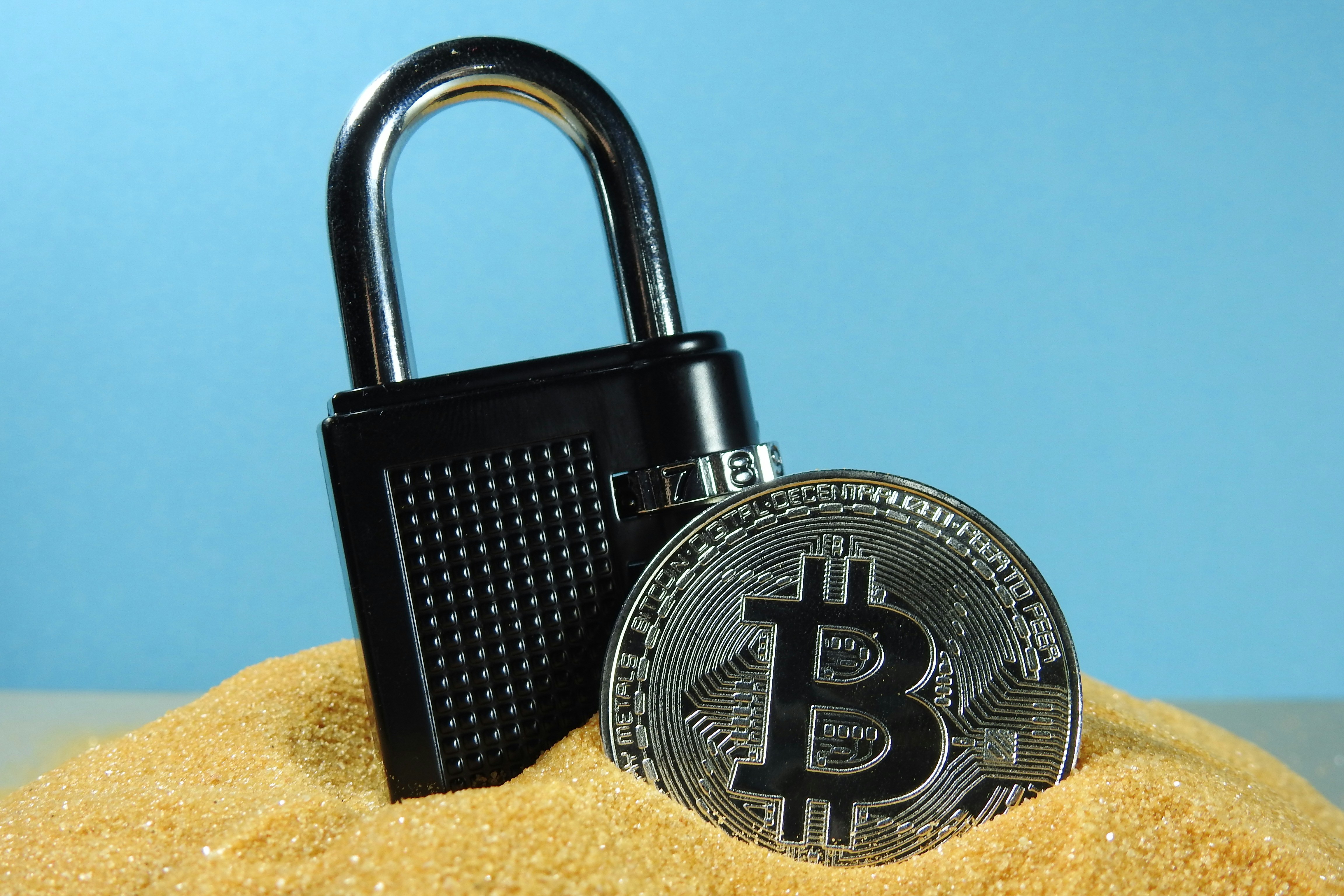 A bitcoin cryptocurrency coin in sand with a black locked padlock
