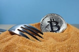 A silver fork partially buried in a mound of brown sugar beside a silver Bitcoin coin against a blue background.