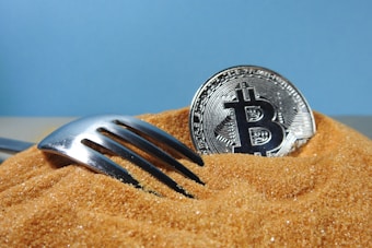 A silver fork partially buried in a mound of brown sugar beside a silver Bitcoin coin against a blue background.