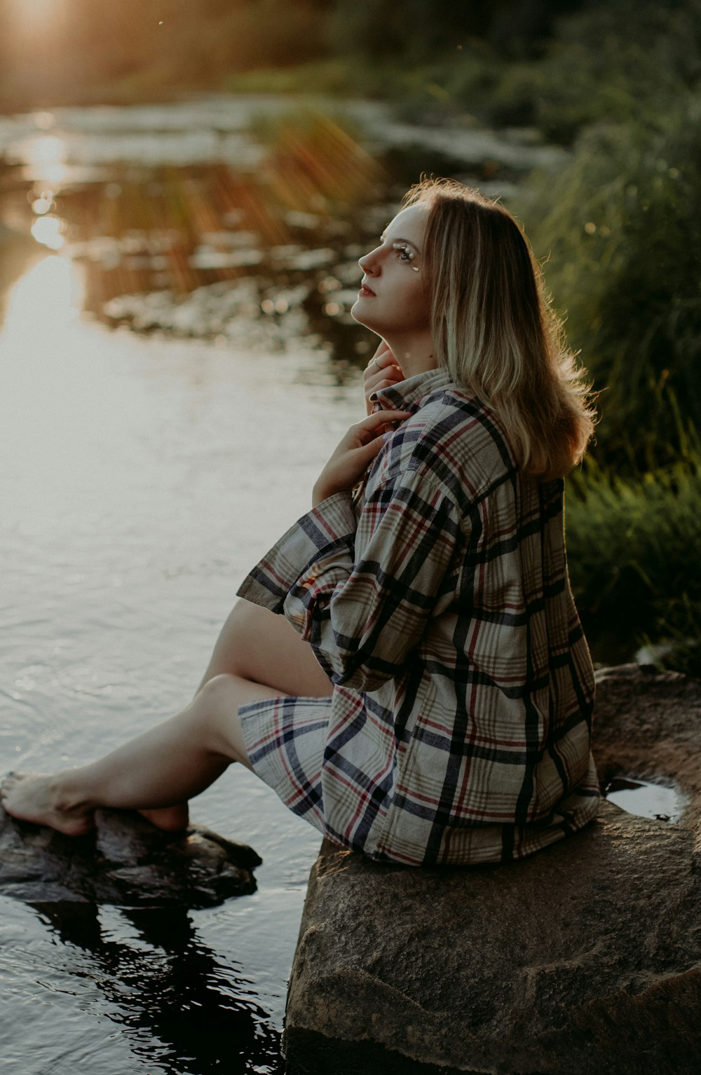 woman in black and white plaid dress shirt sitting on rock near river during daytime