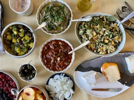 A variety of dishes are arranged on a table. There are bowls of roasted vegetables, a savory stew, a fresh salad with herbs, and a red sauce with visible pieces of tomato. Fresh fruits like peaches and berries are also present, along with a platter of cheese and sliced onions. A small pitcher of olive oil and a container of salt can be seen alongside cutlery.