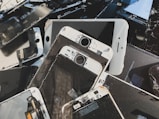 A collection of disassembled and broken smartphone screens and components, primarily displaying the front side of various devices. The parts are scattered in a disorderly manner, displaying signs of wear and damage, including cracked screens and exposed internal circuitry.