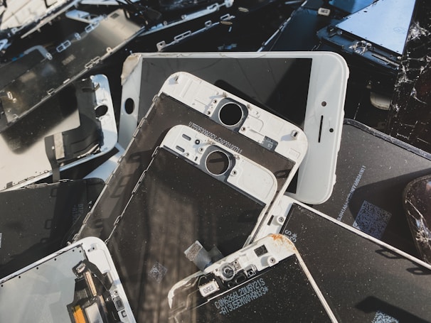 A collection of disassembled and broken smartphone screens and components, primarily displaying the front side of various devices. The parts are scattered in a disorderly manner, displaying signs of wear and damage, including cracked screens and exposed internal circuitry.