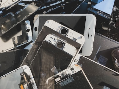 A collection of disassembled and broken smartphone screens and components, primarily displaying the front side of various devices. The parts are scattered in a disorderly manner, displaying signs of wear and damage, including cracked screens and exposed internal circuitry.