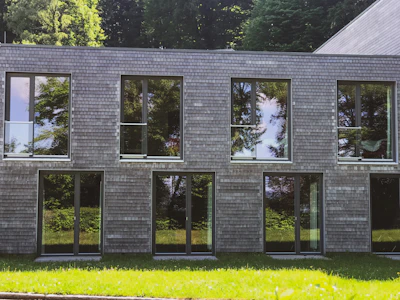 A modern building with a facade made of small, square, wooden shingles. The large windows reflect the lush green trees surrounding the area. The structure has a rectangular shape and the grass in the foreground adds a natural contrast to the shingled exterior.