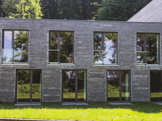 A modern building with a facade made of small, square, wooden shingles. The large windows reflect the lush green trees surrounding the area. The structure has a rectangular shape and the grass in the foreground adds a natural contrast to the shingled exterior.