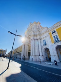 An architectural scene featuring a grand historic arch and building, with ornate details and tall columns, under a bright blue sky. The sun is partially visible, casting a glow and lens flare. The street is lined with people walking and bicycling, and there are modern street lamps and cobblestone paving.
