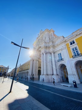 An architectural scene featuring a grand historic arch and building, with ornate details and tall columns, under a bright blue sky. The sun is partially visible, casting a glow and lens flare. The street is lined with people walking and bicycling, and there are modern street lamps and cobblestone paving.
