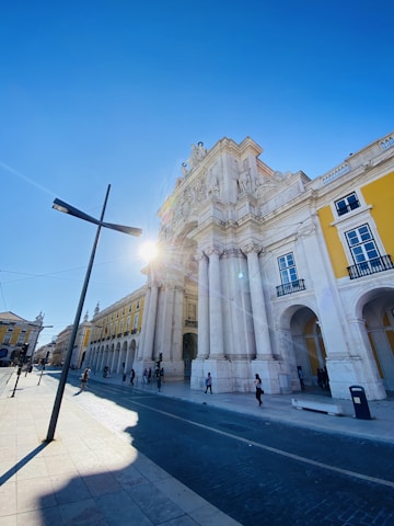 An architectural scene featuring a grand historic arch and building, with ornate details and tall columns, under a bright blue sky. The sun is partially visible, casting a glow and lens flare. The street is lined with people walking and bicycling, and there are modern street lamps and cobblestone paving.