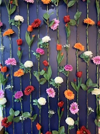 A vertical arrangement of various artificial flowers and leaves attached to a dark blue wall. The flowers include red tulips, orange and white peonies, and pink and red daisies. The stems are aligned in parallel rows, creating an orderly pattern.