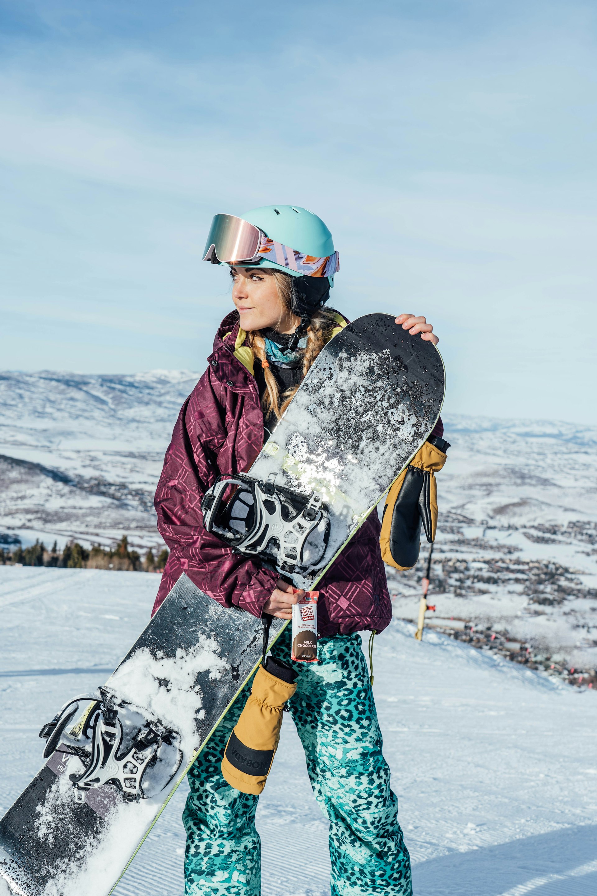 Female snowboarder with helmet and goggles holding her board, mountain backdrop
