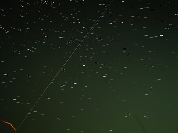 A long exposure shot of the night sky with star trails creating streaks of light. The image features a slightly greenish tint, enhancing the celestial background. There is a distinct line crossing diagonally across the image, perhaps a satellite or aircraft trail, and a faint red streak at the bottom left.