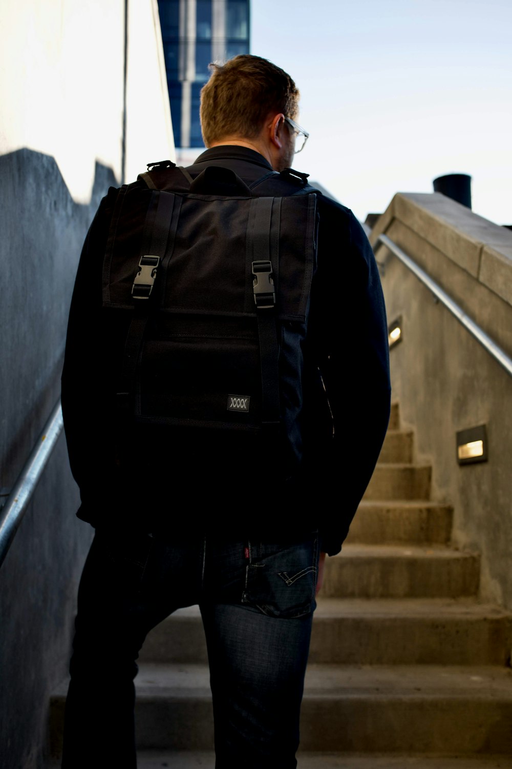 man in black jacket and blue denim jeans with black backpack standing on stairs during daytime