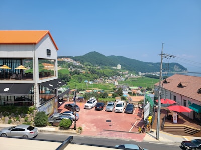 A scenic view featuring a parking lot with multiple cars, a modern building with a cafe or restaurant having outdoor seating, and a picturesque landscape in the background with green hills and a small town. A clear blue sky enhances the serene atmosphere.