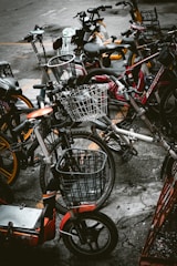 A cluster of various bicycles and e-bikes parked haphazardly on a concrete pavement. The frames of the bikes include a mix of colors such as red, orange, and dark hues, with some equipped with woven wire baskets. The setting is urban, suggesting a communal or shared bike system.