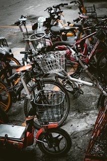 A cluster of various bicycles and e-bikes parked haphazardly on a concrete pavement. The frames of the bikes include a mix of colors such as red, orange, and dark hues, with some equipped with woven wire baskets. The setting is urban, suggesting a communal or shared bike system.