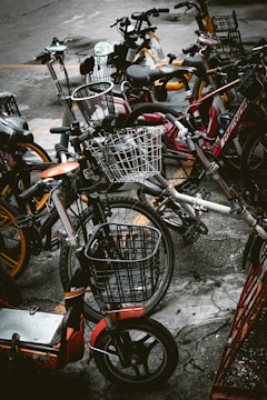 A cluster of various bicycles and e-bikes parked haphazardly on a concrete pavement. The frames of the bikes include a mix of colors such as red, orange, and dark hues, with some equipped with woven wire baskets. The setting is urban, suggesting a communal or shared bike system.