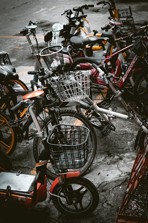 A cluster of various bicycles and e-bikes parked haphazardly on a concrete pavement. The frames of the bikes include a mix of colors such as red, orange, and dark hues, with some equipped with woven wire baskets. The setting is urban, suggesting a communal or shared bike system.