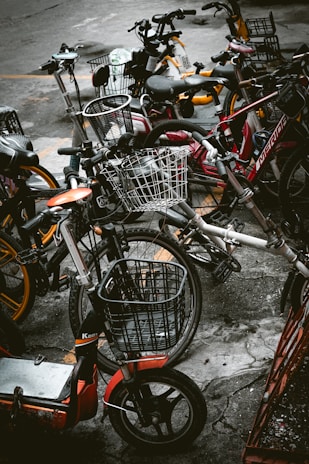 A cluster of various bicycles and e-bikes parked haphazardly on a concrete pavement. The frames of the bikes include a mix of colors such as red, orange, and dark hues, with some equipped with woven wire baskets. The setting is urban, suggesting a communal or shared bike system.