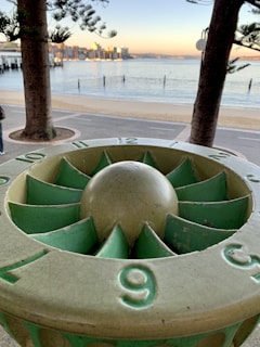 A sundial is prominently positioned in the foreground with surrounding trees framing a view of a calm beach and a distant city skyline. The scene features a clear, calm body of water reflecting soft morning or evening light.