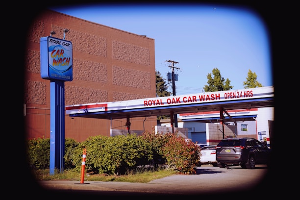 A car wash facility with a prominent blue sign displaying 'Royal Oak Car Wash' beside a drive-through area. The sign also indicates that the car wash is open 24 hours. Multiple cars are parked in the vicinity, and there is a small green hedge along the driveway. A tall building and trees can be seen in the background, with utility poles and wires overhead.