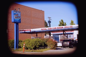 A car wash facility with a prominent blue sign displaying 'Royal Oak Car Wash' beside a drive-through area. The sign also indicates that the car wash is open 24 hours. Multiple cars are parked in the vicinity, and there is a small green hedge along the driveway. A tall building and trees can be seen in the background, with utility poles and wires overhead.
