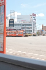 A cityscape featuring a brightly colored orange tram passing through an urban area. In the foreground, there is a bus stop sign with visible schedules. The background includes several buildings with signage, power lines, and a clear blue sky with a few clouds.