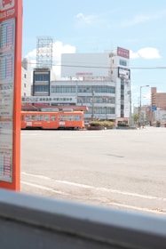 A cityscape featuring a brightly colored orange tram passing through an urban area. In the foreground, there is a bus stop sign with visible schedules. The background includes several buildings with signage, power lines, and a clear blue sky with a few clouds.