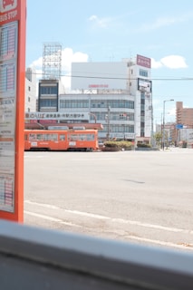 A cityscape featuring a brightly colored orange tram passing through an urban area. In the foreground, there is a bus stop sign with visible schedules. The background includes several buildings with signage, power lines, and a clear blue sky with a few clouds.