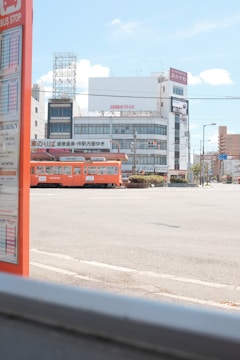 A cityscape featuring a brightly colored orange tram passing through an urban area. In the foreground, there is a bus stop sign with visible schedules. The background includes several buildings with signage, power lines, and a clear blue sky with a few clouds.