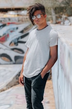 A person with dreadlocks and glasses leans casually against a graffiti-covered wall in an outdoor urban setting. The person is wearing a black and white striped shirt and black pants. The scene conveys a laid-back, artistic vibe.