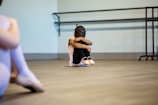 A young girl is sitting on the floor of a dance studio, with her arms wrapped around her knees and head tucked down. She wears ballet attire, including ballet shoes, and her hair is styled in braids. The background includes a barre mounted on the wall. Another person, out of focus, is partially visible on the left side of the image, also dressed in ballet clothes.