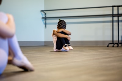 A young girl is sitting on the floor of a dance studio, with her arms wrapped around her knees and head tucked down. She wears ballet attire, including ballet shoes, and her hair is styled in braids. The background includes a barre mounted on the wall. Another person, out of focus, is partially visible on the left side of the image, also dressed in ballet clothes.