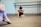 A young girl is sitting on the floor of a dance studio, with her arms wrapped around her knees and head tucked down. She wears ballet attire, including ballet shoes, and her hair is styled in braids. The background includes a barre mounted on the wall. Another person, out of focus, is partially visible on the left side of the image, also dressed in ballet clothes.