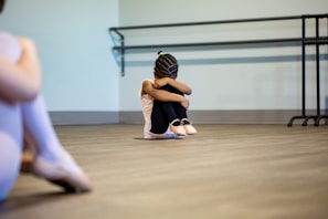 A young girl is sitting on the floor of a dance studio, with her arms wrapped around her knees and head tucked down. She wears ballet attire, including ballet shoes, and her hair is styled in braids. The background includes a barre mounted on the wall. Another person, out of focus, is partially visible on the left side of the image, also dressed in ballet clothes.