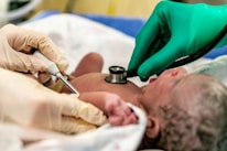 A newborn baby is being examined by medical professionals. One person is using an ultrasound device while another uses a stethoscope to check the baby's health. The baby lies on a white cloth, surrounded by individuals wearing gloves, emphasizing a clinical setting.