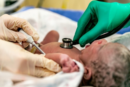 A newborn baby is being examined by medical professionals. One person is using an ultrasound device while another uses a stethoscope to check the baby's health. The baby lies on a white cloth, surrounded by individuals wearing gloves, emphasizing a clinical setting.
