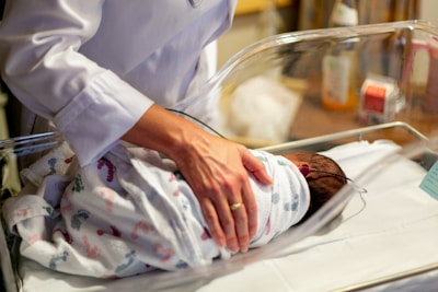 A newborn baby is gently wrapped in a patterned blanket, lying in a clear hospital bassinet. A person wearing a white coat, possibly a healthcare professional, is placing their hand on the baby, indicating care and attention. The background shows some medical equipment and supplies, signifying a hospital setting.