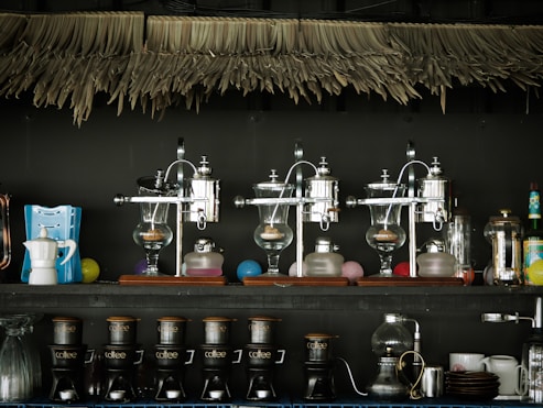 A collection of coffee-making equipment is displayed on shelves. The upper shelf holds three vintage-style coffee siphons and various decorative items. The lower shelf has multiple coffee drippers labeled with 'coffee,' along with a moka pot, cups, and other utensils. Above the shelves, there is a decorative fringed canopy.