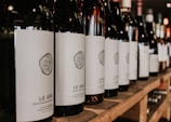 A row of wine bottles is neatly arranged on a wooden shelf, with each bottle featuring a minimalist white label and a stylized logo. The focus is on the bottles in the foreground, while additional bottles blur into the background, suggesting a well-stocked collection.