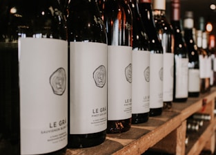A row of wine bottles is neatly arranged on a wooden shelf, with each bottle featuring a minimalist white label and a stylized logo. The focus is on the bottles in the foreground, while additional bottles blur into the background, suggesting a well-stocked collection.